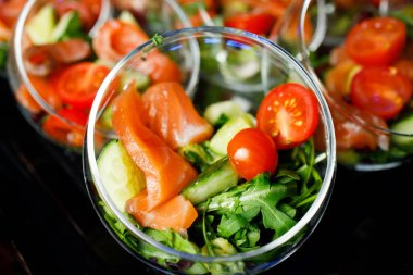Üzerinde kırmızı balık salatası, domates, salatalık ve bitkilerden oluşan cam bardaklar olan açık büfe masasının üstünde.