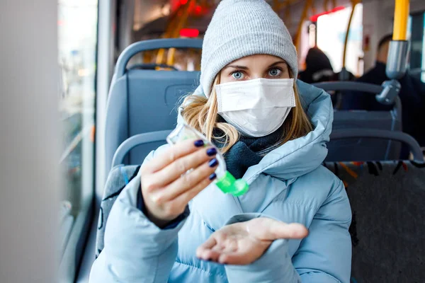 Блондинка в медицинской маске, глядя в сторону, дезинфицирует руки инструментом, сидя в автобусе возле окна. — стоковое фото