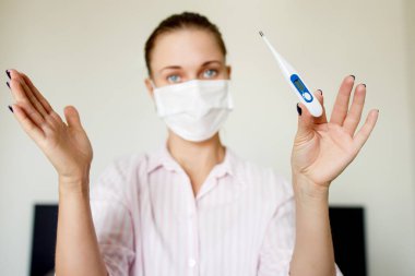 Evinde elinde termometre olan tıbbi maskeli bir kadın..
