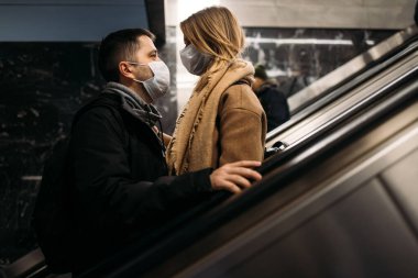 Genç adam ve sarışın kadın metroda yürüyen merdivende tıbbi maskeler takıp birbirlerine bakıyorlar..
