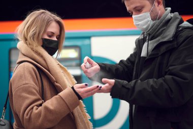 Tıbbi maskeli genç adam ve sarışın kadın yeraltı vagonunun yanında dururken antiseptik kullanıyor..