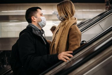 Metronun yürüyen merdivenlerinde birbirlerine tıbbi maskelerle bakan sevgi dolu bir kadın ve erkek..