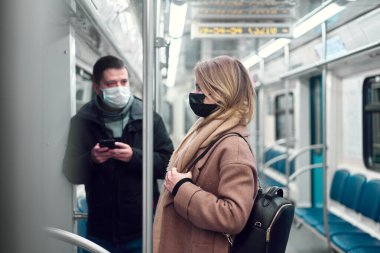 Elinde telefon olan esmer bir adam ve metro vagonunda duran maskeli sarışın bir kadın..