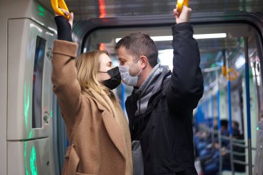 Tıbbi maskeli genç bir çift metro vagonunda tırabzan tutuyor..