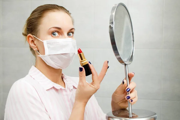 립스틱 과 거울을 손에 들고 의료용 마스크를 쓴 금발 소녀의 근접 사진 — 스톡 사진