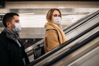 Tıbbi maskeli genç adam ve kadının metroda yürüyen merdivende yan görüntüsü..