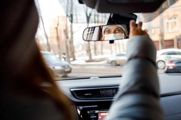 Женщина в медицинской маске днем сидит за рулем в машине. Отражение в зеркале
