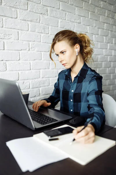 Mujer rubia freelancer sentado en la mesa con el ordenador portátil en el fondo de la pared de ladrillo blanco . — Foto de Stock