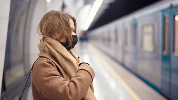 Vista lateral de la joven rubia con máscara médica negra parada en el metro cerca del carruaje . — Foto de Stock