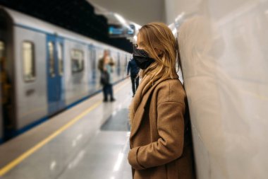 Siyah tıbbi maskeli genç sarışın kadın metro vagonunun yanında duruyor. - Evet. Yan görünüm