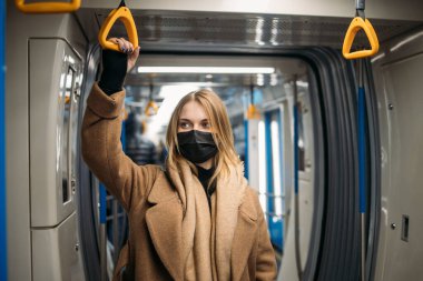 Siyah tıbbi maskeli kadın metroda sarı korkuluklara tutunuyor..