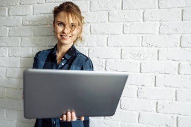 Elinde dizüstü bilgisayarla beyaz tuğla duvara karşı duran mutlu bir serbest çalışan kadın..