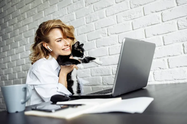 Smiling woman with black cat in her arms sitting at table with laptop.