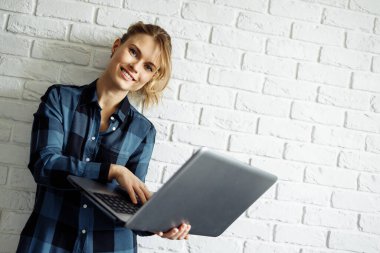 Elinde dizüstü bilgisayarla beyaz tuğla duvara yaslanmış gülümseyen sarışın serbest yazar.