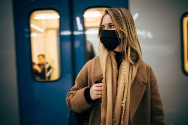 Siyah tıbbi maskeli sarışın kadın metroda arabanın yanında dikilirken yan bakıyor.