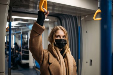 Siyah tıbbi maskeli sarışın metroda sarı korkuluklara tutunuyor.