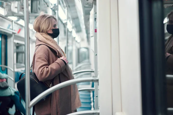 Wanita berambut pirang bertopeng medis hitam berdiri di kereta bawah tanah. — Stok Foto