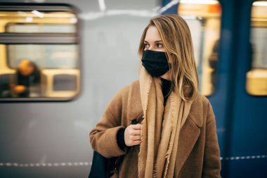 Siyah tıbbi maskeli genç sarışın metroda arabanın yanında dikilirken yan bakıyor..