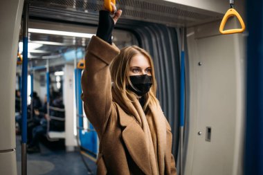 Siyah tıbbi maskeli genç bir kadın metroda sarı korkuluklara tutunuyor..
