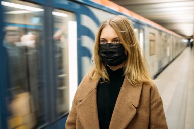 Siyah tıbbi maskeli genç bir kadın metro vagonunun yanında kameraya bakıyor..