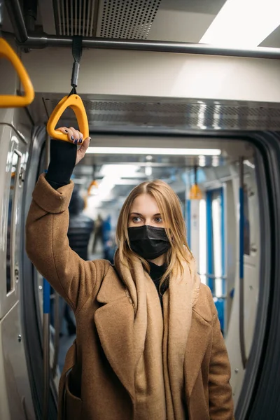 Siyah tıbbi maskeli sarışın kadın metroda sarı korkuluklara tutunuyor..
