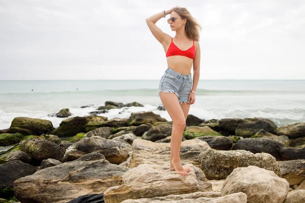 Jovem mulher de fato de banho vermelho e calções jeans em pé sobre pedras — Fotografia de Stock