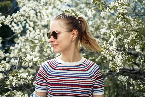 Молодая женщина в солнечных очках смотрит в сторону, стоя на фоне цветущей яблони — стоковое фото
