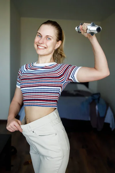 Mujer delgada sonriente en pantalones grandes con mancuerna en la mano en el apartamento . —  Fotos de Stock