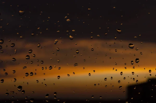 Lluvia fuera de la ventana en el fondo de la puesta del sol. Gotas de lluvia sobre vidrio durante la lluvia. Textura brillante de gotas de agua — Foto de Stock