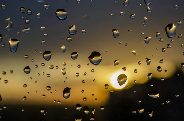 Lluvia fuera de la ventana en el fondo de la puesta del sol. Gotas de lluvia sobre vidrio durante la lluvia. Textura brillante de gotas de agua — Foto de Stock