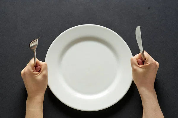 Fork and knife in hands on black background with white plate — 图库照片