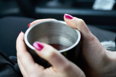 Genç bir kadın arabanın bagajında oturuyor ve elinde bir fincan sıcak içecek tutuyor.