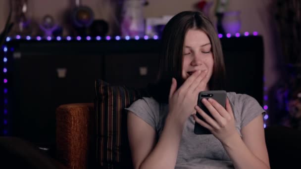 Schöne junge Frau, die zu Hause auf der Couch sitzt und lächelnd auf das Telefon schaut und eine Nachricht tippt — Stockvideo
