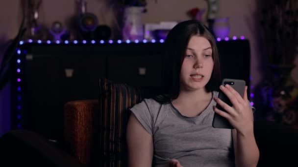 Genç, mutlu, beyaz bir kadın arkadaşlarıyla görüntülü sohbet için cep telefonu kullanıyor ve rahat bir evde otururken sosyal medya için blog yazıyor. — Stok video