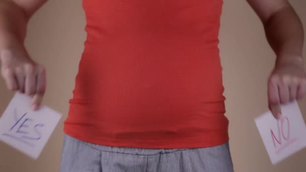 Uma visão de perto da barriga de uma mulher grávida em uma camiseta vermelha, que segura em suas mãos dois lençóis com as palavras SIM e NÃO. A menina escolhe NÃO. Tomar decisões durante a gravidez . — Vídeo de Stock