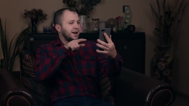 Avrupalı görünümlü genç bir adam ekose gömlekli bir sandalyede oturuyor ve video bağlantısıyla iletişim kuruyor. Abonelerin beğenmesini istiyor. Sosyal ağ blogları kavramı. — Stok video