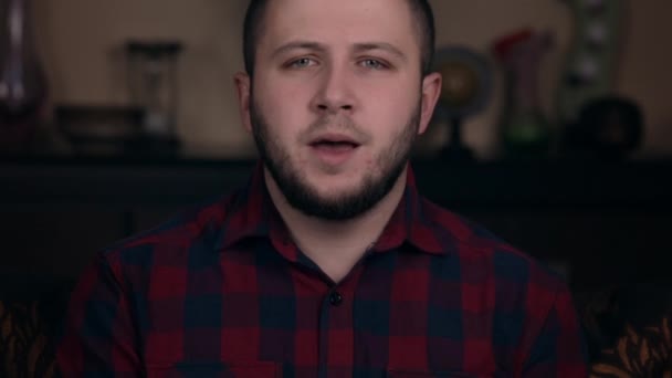 Un jeune homme d'apparence européenne s'assoit sur une chaise dans une chemise à carreaux bouche chaude, répétant un discours avant d'enregistrer un vlog — Video