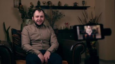 Avrupalı görünümlü bir blogcu olan etkili Sakallı Genç, bir sandalyede tişörtle oturmuş video kaydediyor. Sosyal Medya Günlük Kavramı.