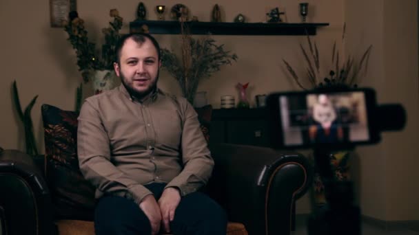 El influyente Barbudo Joven, un blogger, de apariencia europea, se sienta en una silla en una camisa, dispara video en el teléfono y pide que le guste el video. Concepto de Blogging en Redes Sociales — Vídeo de stock