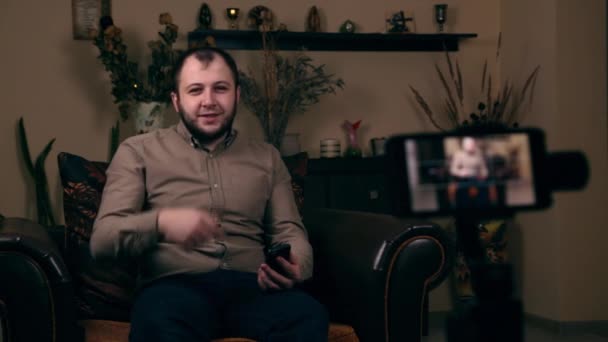 El influyente joven barbudo, un blogger, de apariencia europea, se sienta en una silla, graba un video vlog y responde a las preguntas de sus seguidores. Concepto de Blogging en Redes Sociales — Vídeo de stock
