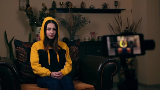 Młoda dziewczyna o europejskim wyglądzie siedzi w fotelu w domu, nagrywa wideo vlog przez telefon i pokazuje kciuk do góry i prosi o to. Koncepcja blogowania mediów społecznościowych — Wideo stockowe