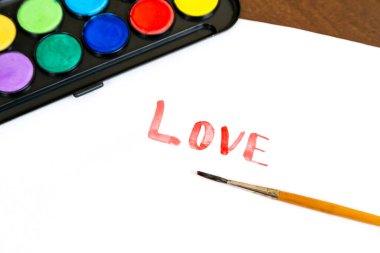 Beyaz arka planda fırça ve suluboya boyalarla el yazısıyla yazılmış bir aşk metni. Resim yapmak için fırçayla boyanmış bir kutu suluboya boya. Aşk konsepti