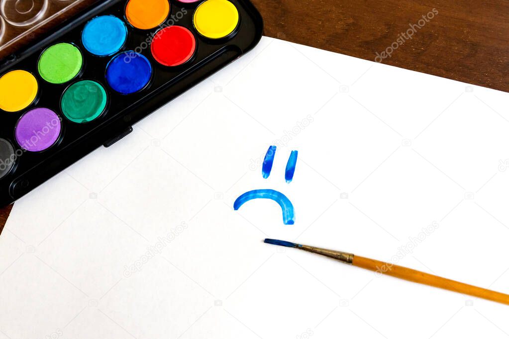 Sad smile face painted with brush and watercolor paints on white background. Box of watercolor paints with a brush for drawing. Emotion concept