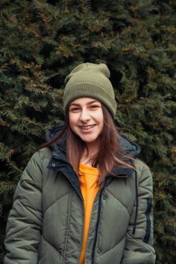 Sarı kazaklı ve haki şapkalı güzel gülümseyen bir kızın dikey portresi. Temiz havada bir Noel ağacının yanında duruyor.