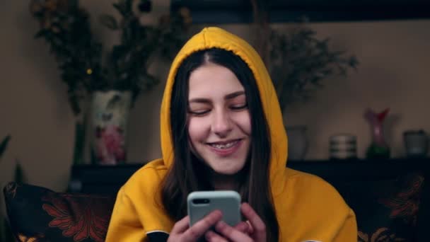Krásná mladá žena sedí na gauči doma s úsměvem při pohledu na telefon a píše zprávu. — Stock video