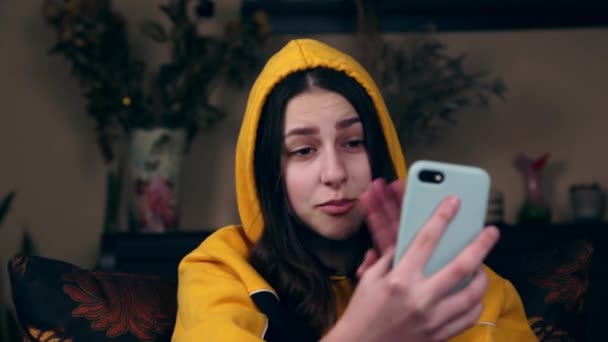 Jeune femme caucasienne heureuse dans une veste jaune en utilisant le téléphone mobile pour le chat vidéo en ligne avec des amis et bloguer pour les médias sociaux tout en étant assis dans la salle à la maison confortable — Video