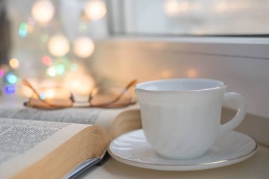 Pencerenin kenarında beyaz bir bardak var, kitabın üzerinde bir kitap yatıyor, gözlük yalan söylüyor.