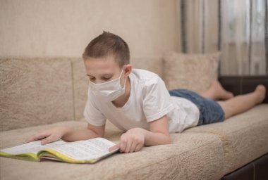 Maske takmış bir çocuk evde kitap okuyor, internetten öğreniyor.