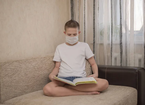 a child in a medical mask reads a book at home, at home, sitting on a sofa, relaxing with a book