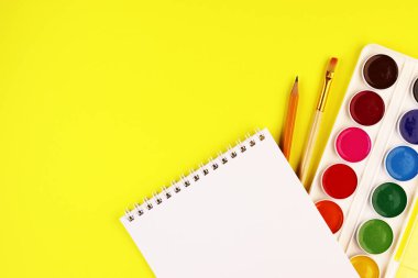 Sarı arka planda okul kırtasiyesi, üst manzara. Boya, not defteri ve renkli kalemler.